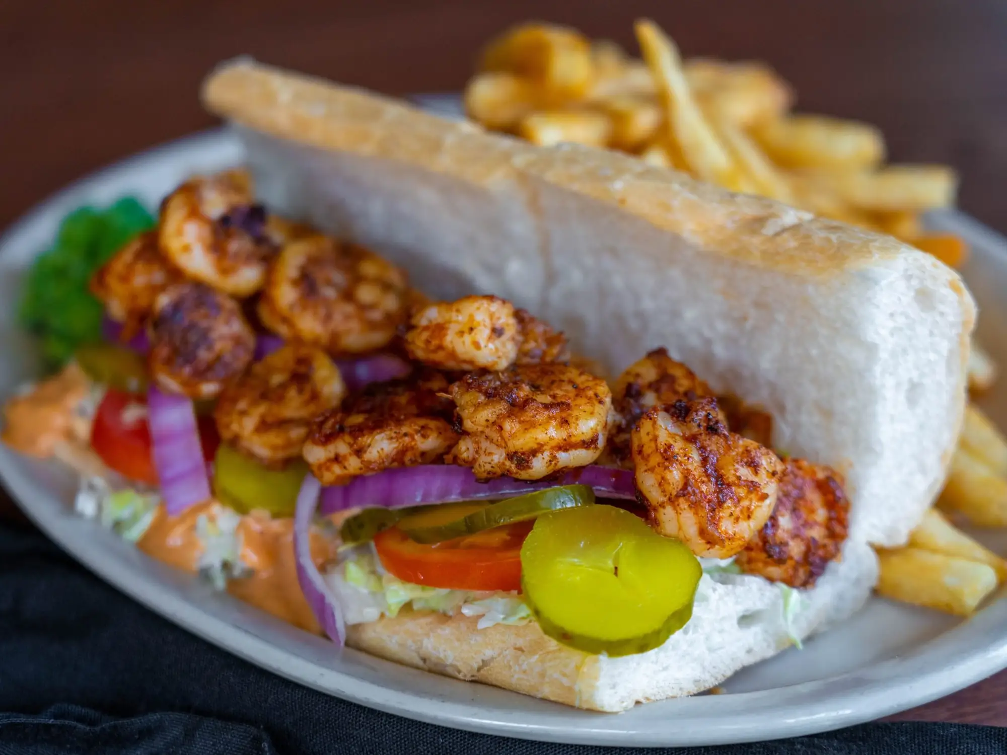 A blackened shrimp po-boy dressed with lettuce, tomato and pickles on French bread, with fries