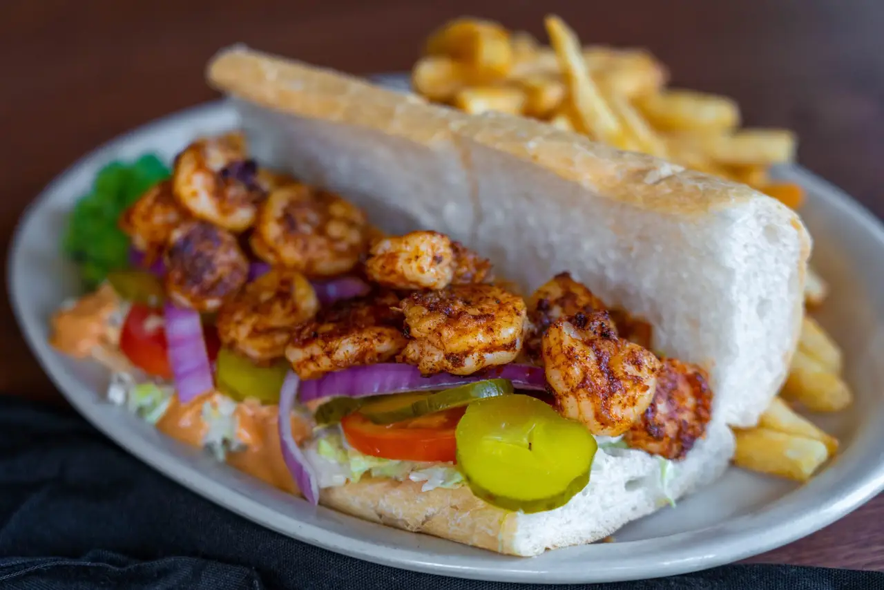 A blackened shrimp po-boy dressed with lettuce, tomato and pickles on French bread, with fries