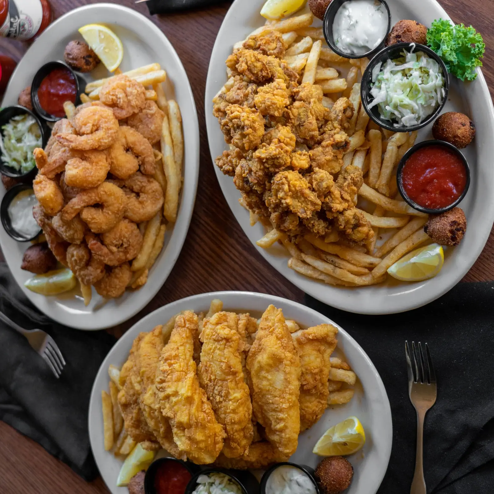 Three fried seafood platters from above: catfish, shrimp and oysters with fries, coleslaw, hushpuppies and sauces
