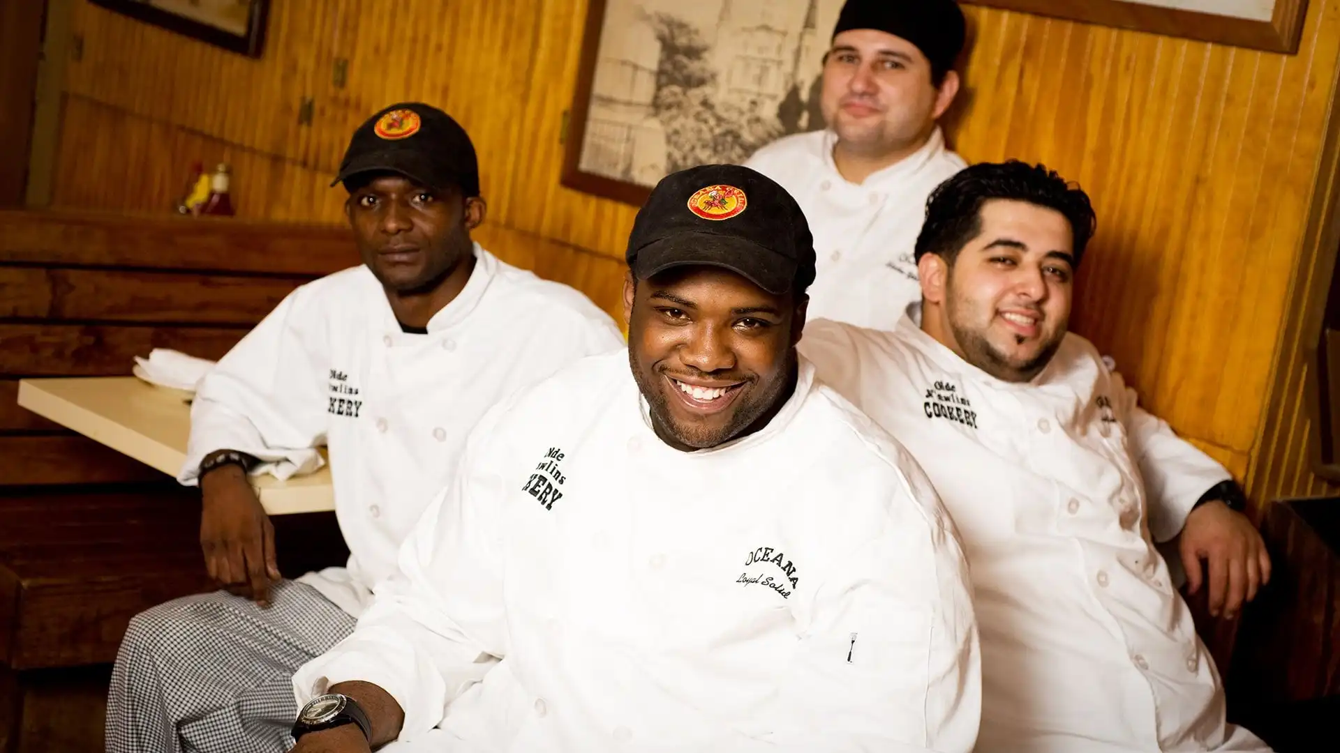 Three Oceana Grill cooks in chef whites and caps, smiling in a booth