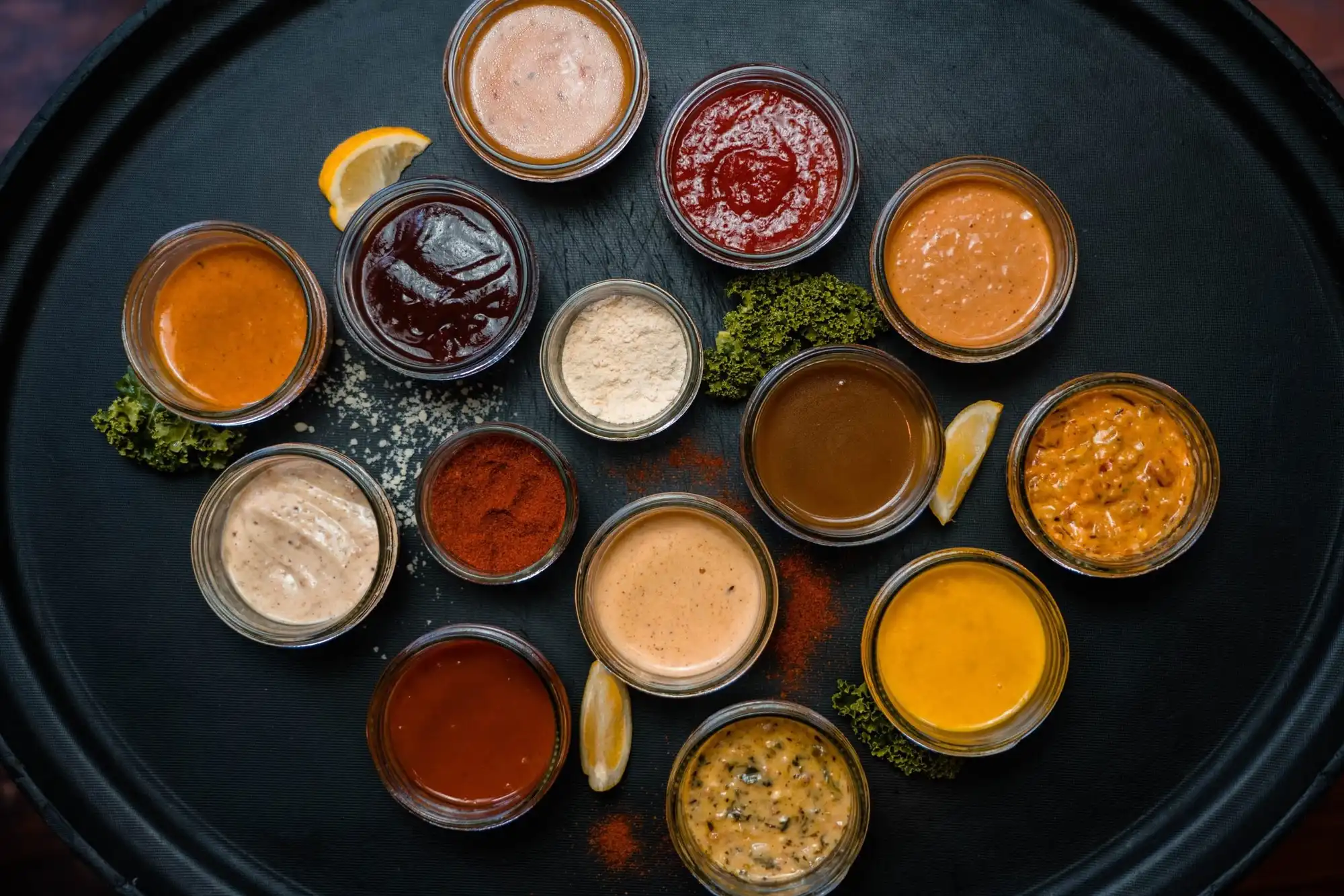 A dozen house sauces and seasonings in ramekins on a black tray, seen from above