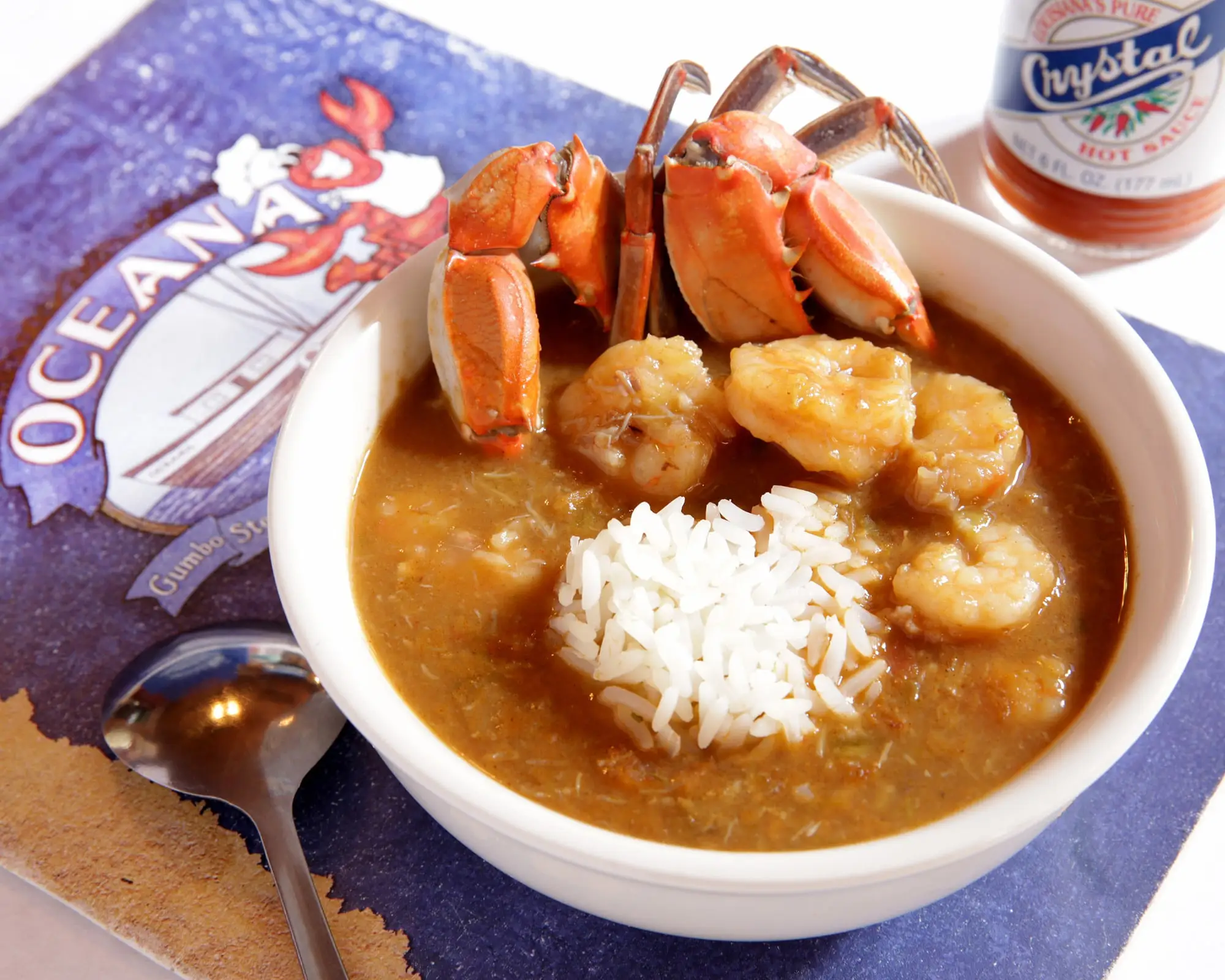 A bowl of seafood gumbo with shrimp, a blue crab and white rice, with Louisiana hot sauce